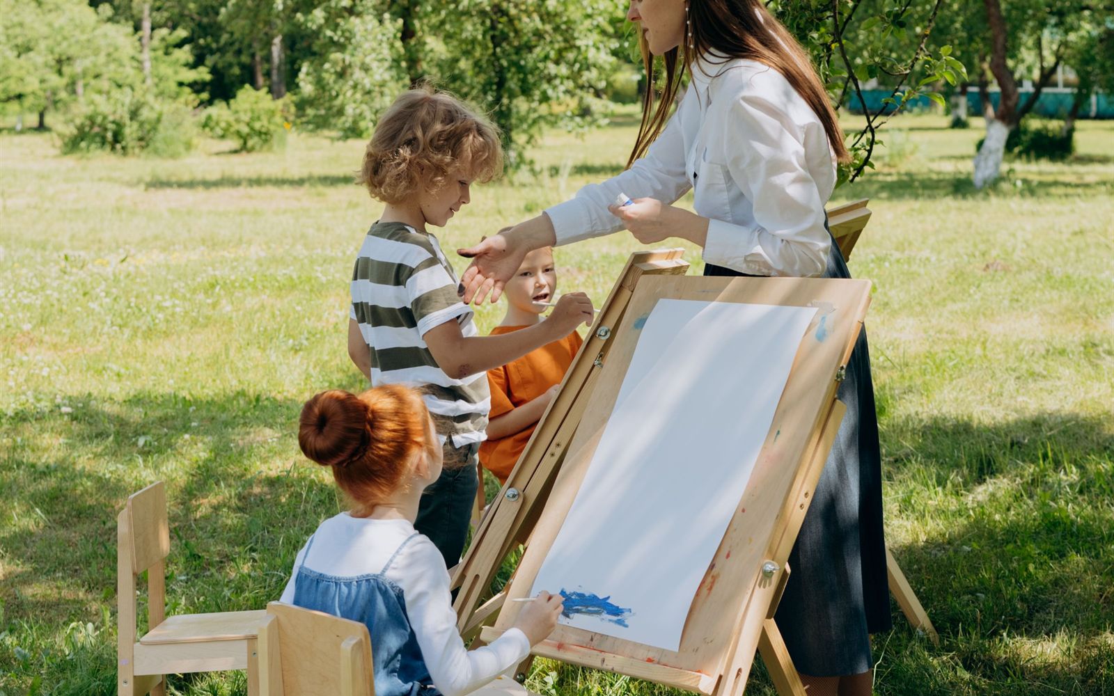 Children painting outside under a tree with their teacher