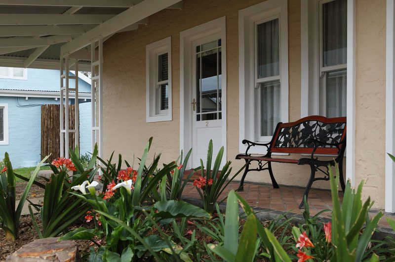 A cottage porch with a wrought-iron bench and flowering plants beside the door