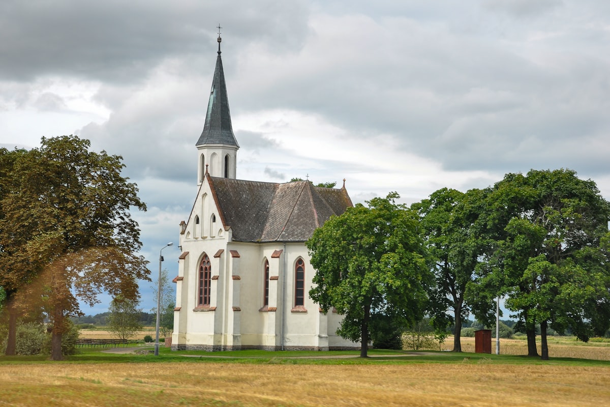 A white chapel with a tall spire, surrounded by trees and open fields